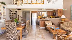 A spacious living area with a dining table, chairs, and a sofa. A kitchen with wooden cabinets and a refrigerator is visible in the background. - Ma'alaea Surf, Unit G10, 2BR Beachfront Condo, AC, Wi-Fi, Pool, TV, Kitchen (Kihei)
