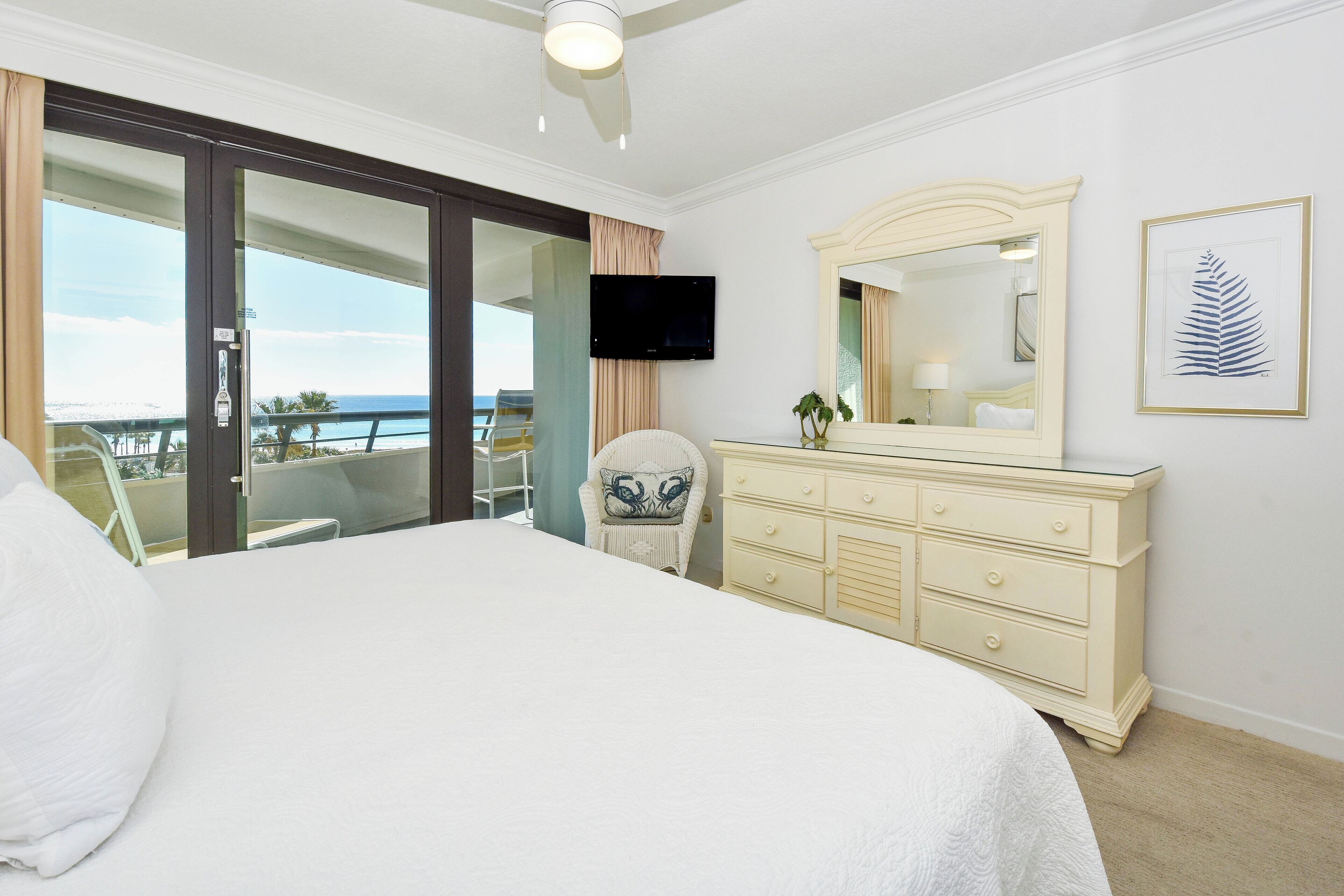 Ein Hotelzimmer mit einem großen Bett, einem Schminktisch, einem Fernseher und einem Balkon mit Blick auf den Ozean.