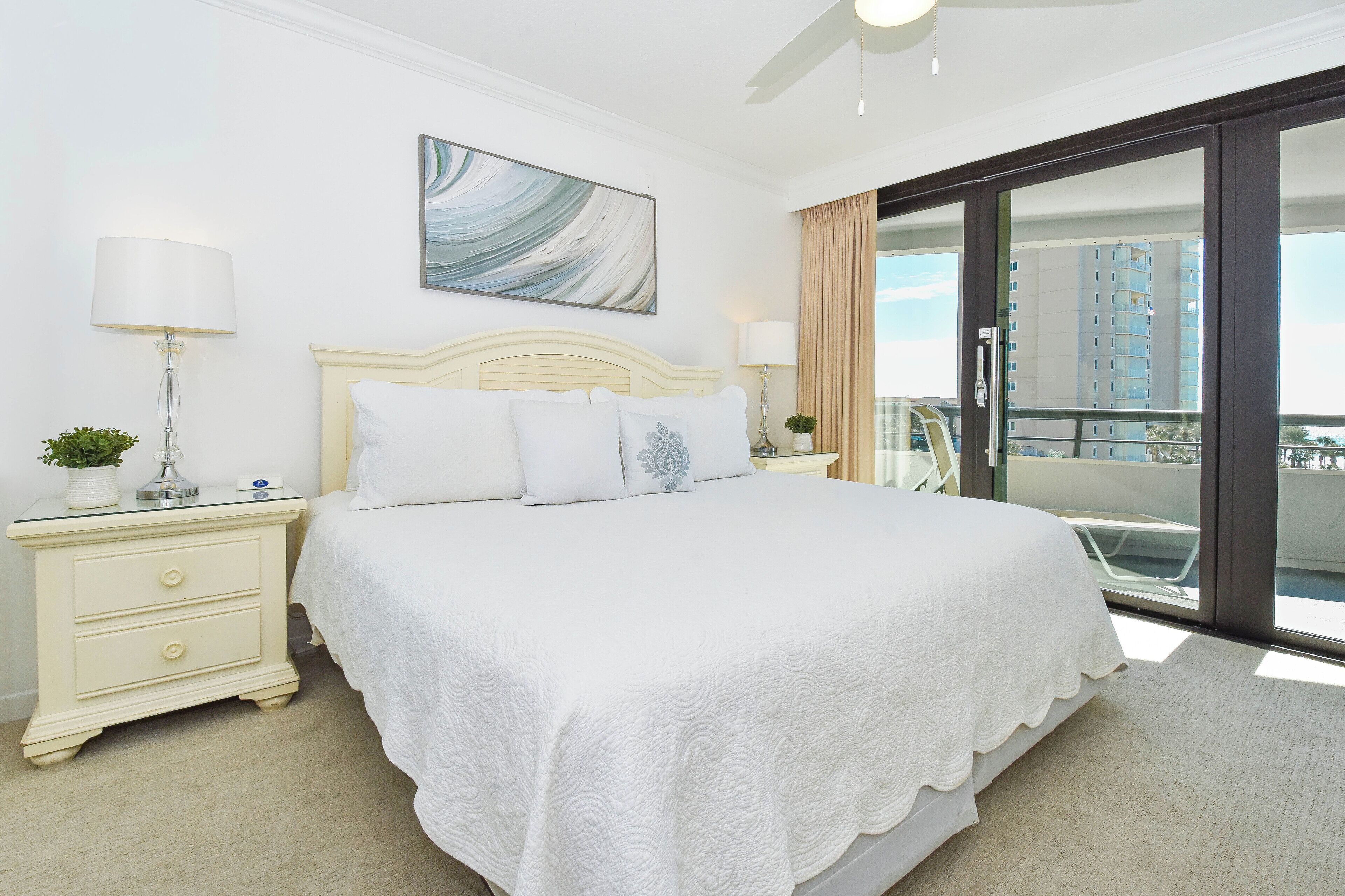 A hotel room with a large bed, a nightstand with a lamp, a painting on the wall, and a view of a building through a glass door.