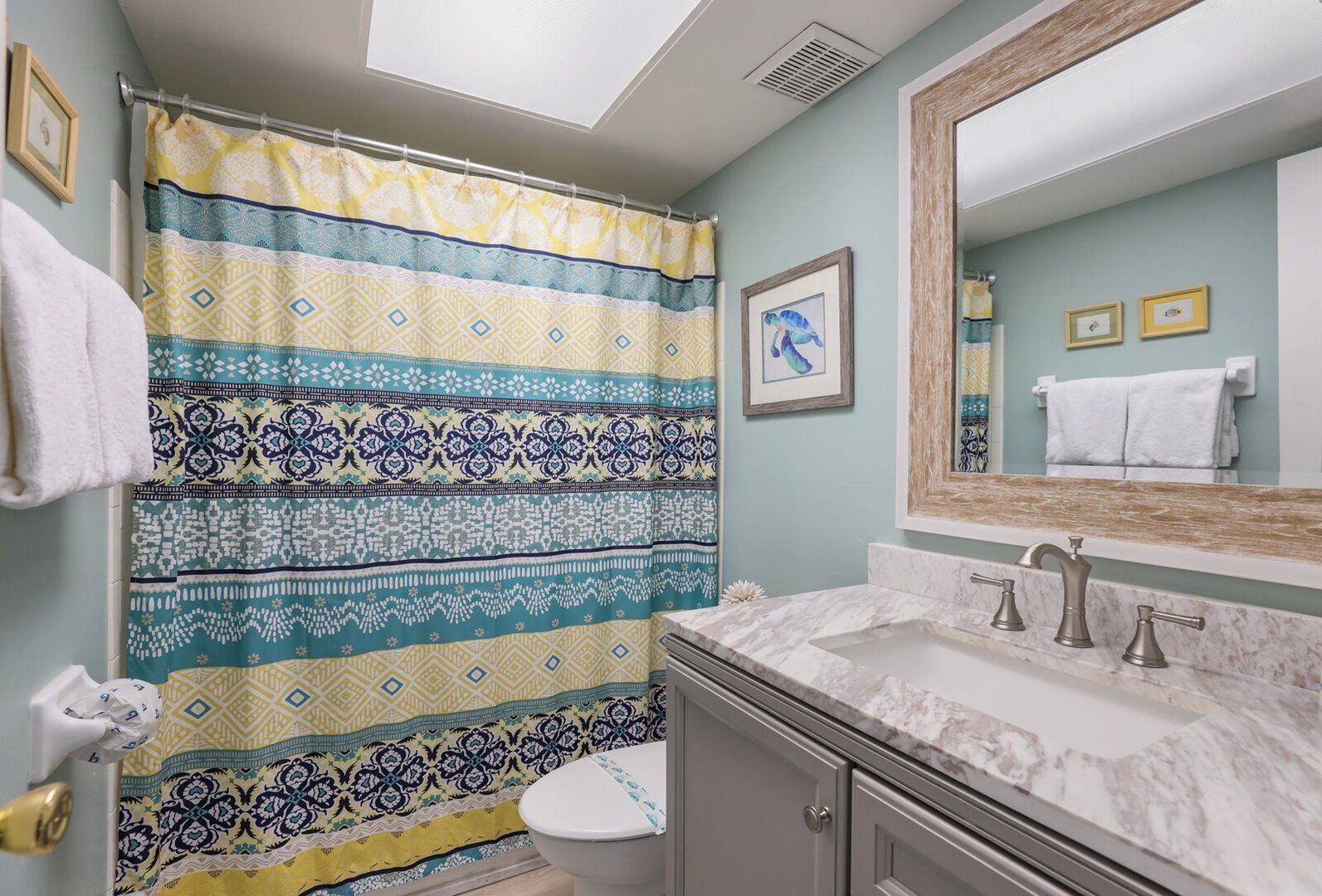 A bathroom with a patterned shower curtain, a toilet, a sink with a marble countertop, and a mirror with a wooden frame.