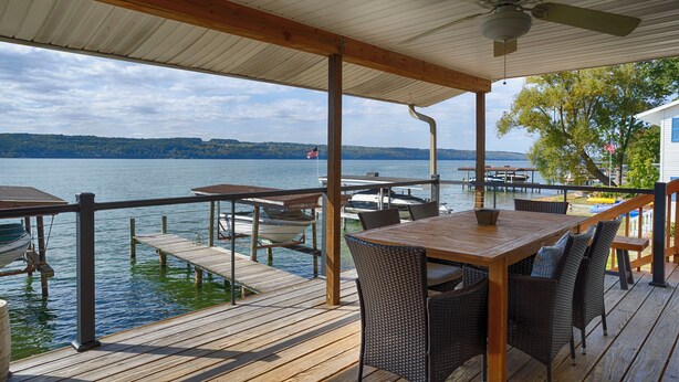 Una terraza de madera con una mesa y sillas, con vista a un lago, barcos amarrados y una orilla lejana.