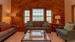 Una sala de estar acogedora con techo de madera, dos ventanas con vistas al exterior, una mesa de centro de vidrio y dos sofás con cojines.