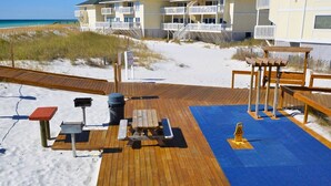 Un'area prendisole in legno con tavoli da picnic e una griglia per barbecue, con vista su una spiaggia e un edificio sullo sfondo.