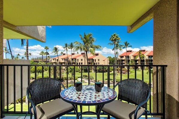 A balcony with two wicker chairs and a small table, overlooking a landscaped area with palm trees and buildings in the background.