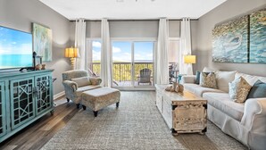 A spacious living room with a large window, a ceiling fan, a sofa, a coffee table, and a cabinet. - Rates Reduced! 2BR Condo with Great Gulf Views! Walk to Beach, Beautiful Pool (Miramar Beach)