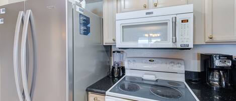 A compact kitchen with a stainless steel refrigerator, a microwave, an electric stove, and a coffee maker.