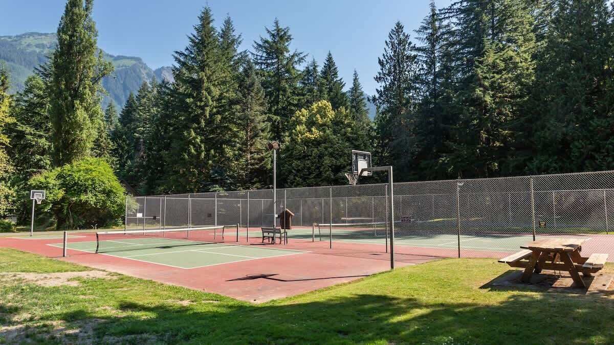 A tennis court with a picnic table and a basketball hoop, surrounded by trees and a mountain backdrop.