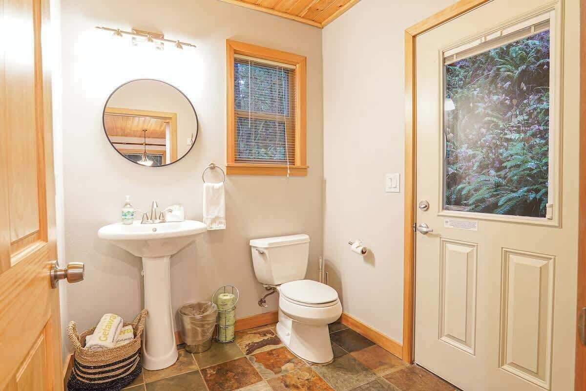 A bathroom with a sink, toilet, mirror, window, and a door with a view of a forest.