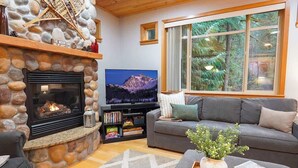Ein gemütliches Wohnzimmer mit Kamin aus Stein, einem großen Flachbildfernseher, einem grauen Sofa und einem Fenster mit Blick auf den Wald.