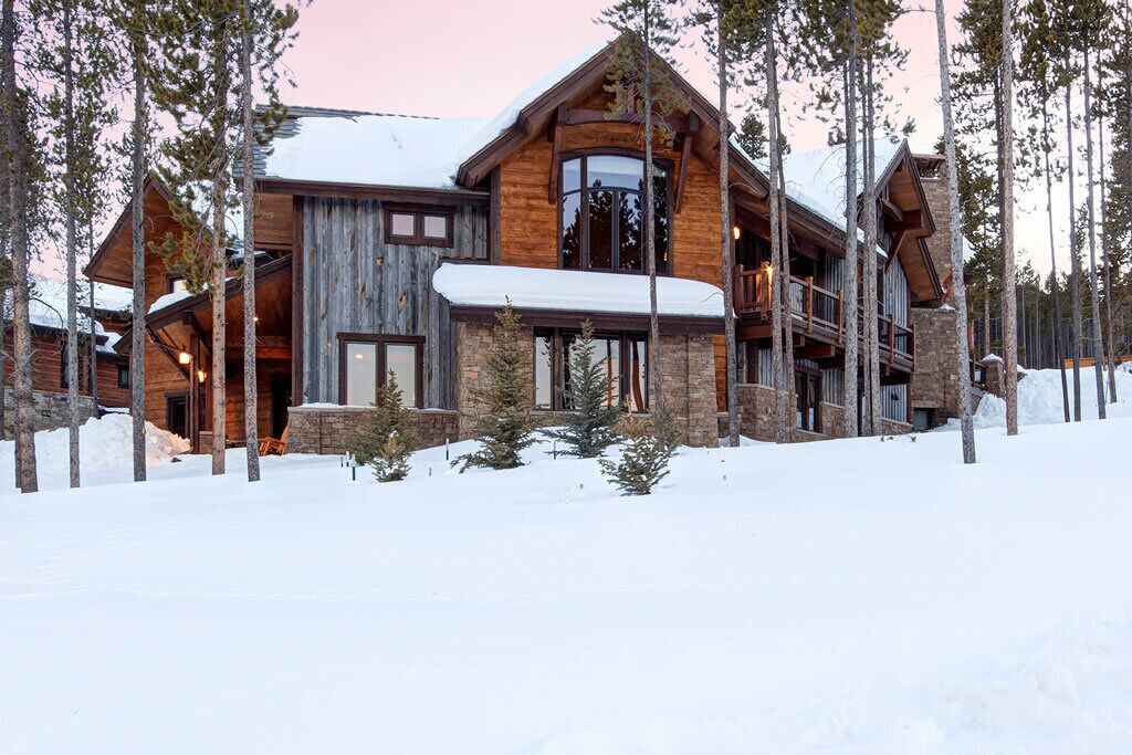 A large log cabin with multiple levels, stone foundations, and a snow-covered landscape.