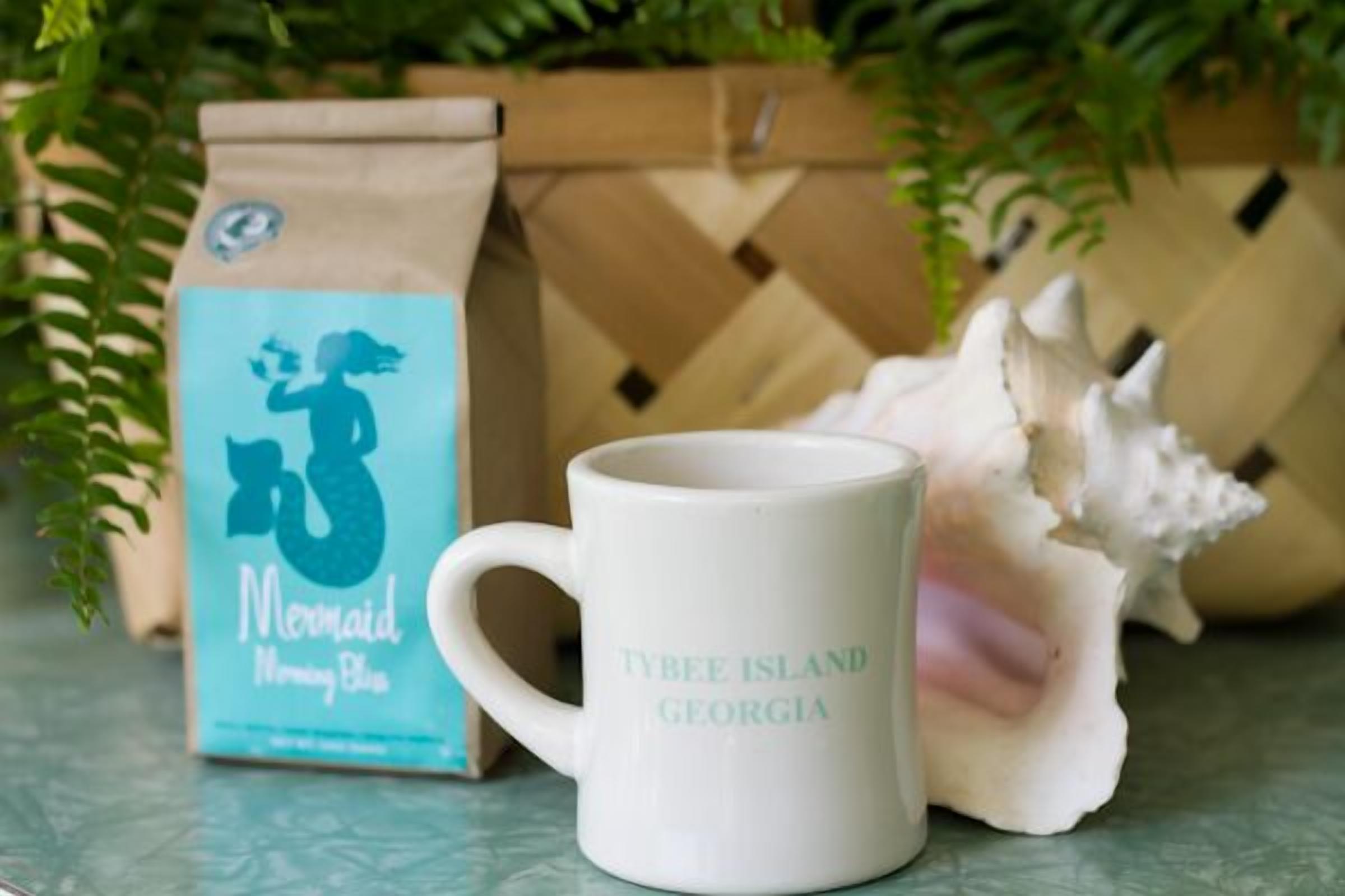A white mug with "Tybee Island Georgia" text, a coffee bag with a mermaid logo, and a seashell decoration.