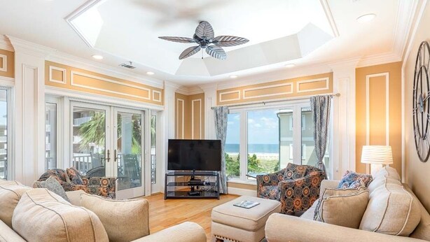 A spacious living room with a ceiling fan, a flat-screen TV, and a large window offering a view of the ocean.