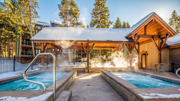 A hot tub area with a wooden deck and a covered seating area, surrounded by trees and a building with a snow-covered roof. - Enchanting Mountain Haven: Ski, Explore & Relax with Unrivaled VIEWS! (Breckenridge)