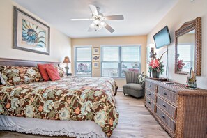 A bedroom with a large bed, a ceiling fan, a dresser, a chair, and a TV on the wall.