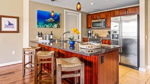 A kitchen with a central island, wooden chairs, a large refrigerator, and a painting of a sea turtle.