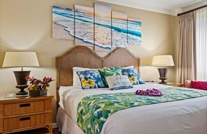 Una habitación de hotel con una cama grande, cabecera de madera, almohadas estampadas con flores, colcha con motivos tropicales y un mural de playa en la pared.