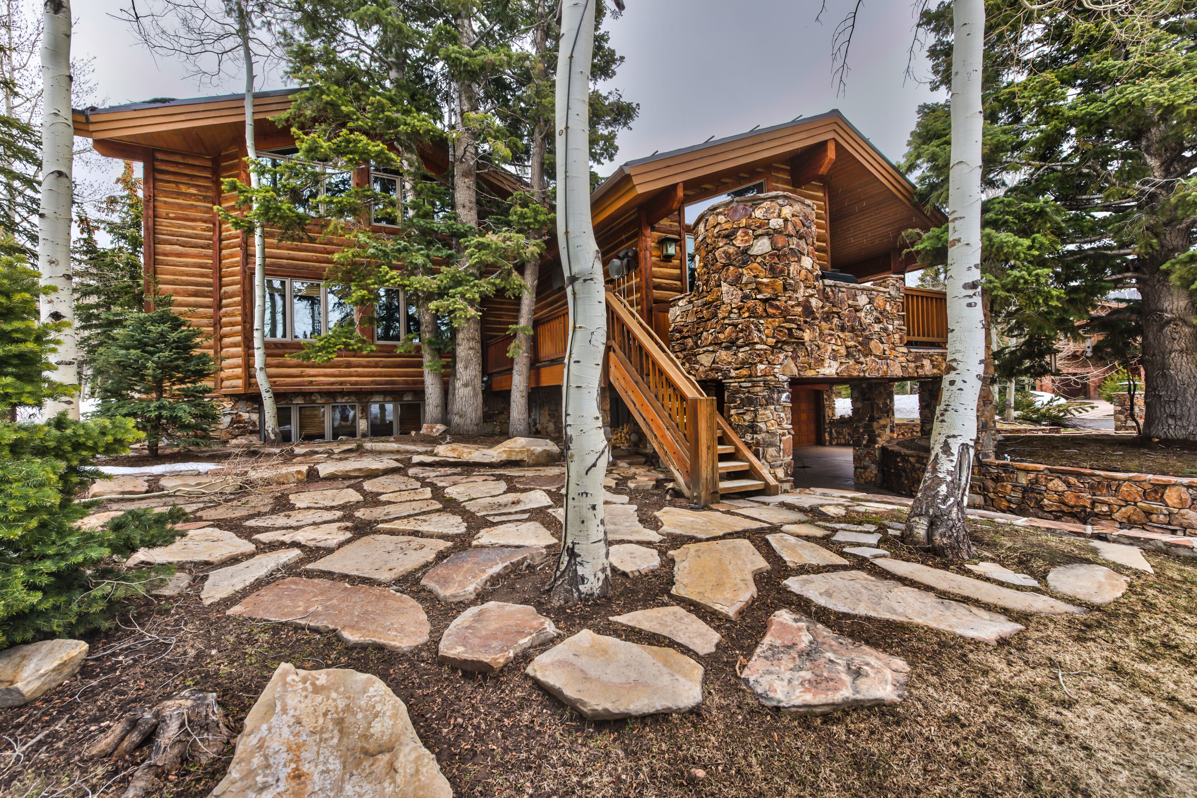 A log cabin with a stone staircase leading to a wooden deck, surrounded by birch trees and a stone pathway.