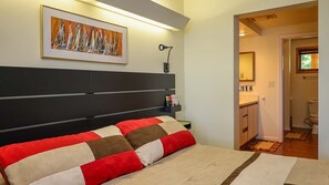 Une chambre avec un grand lit, une tête de lit, une table de chevet, une lampe, un tableau encadré au mur et une vue sur la salle de bain.