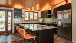 A modern kitchen with a large island, bar stools, a stainless steel refrigerator, and pendant lighting.