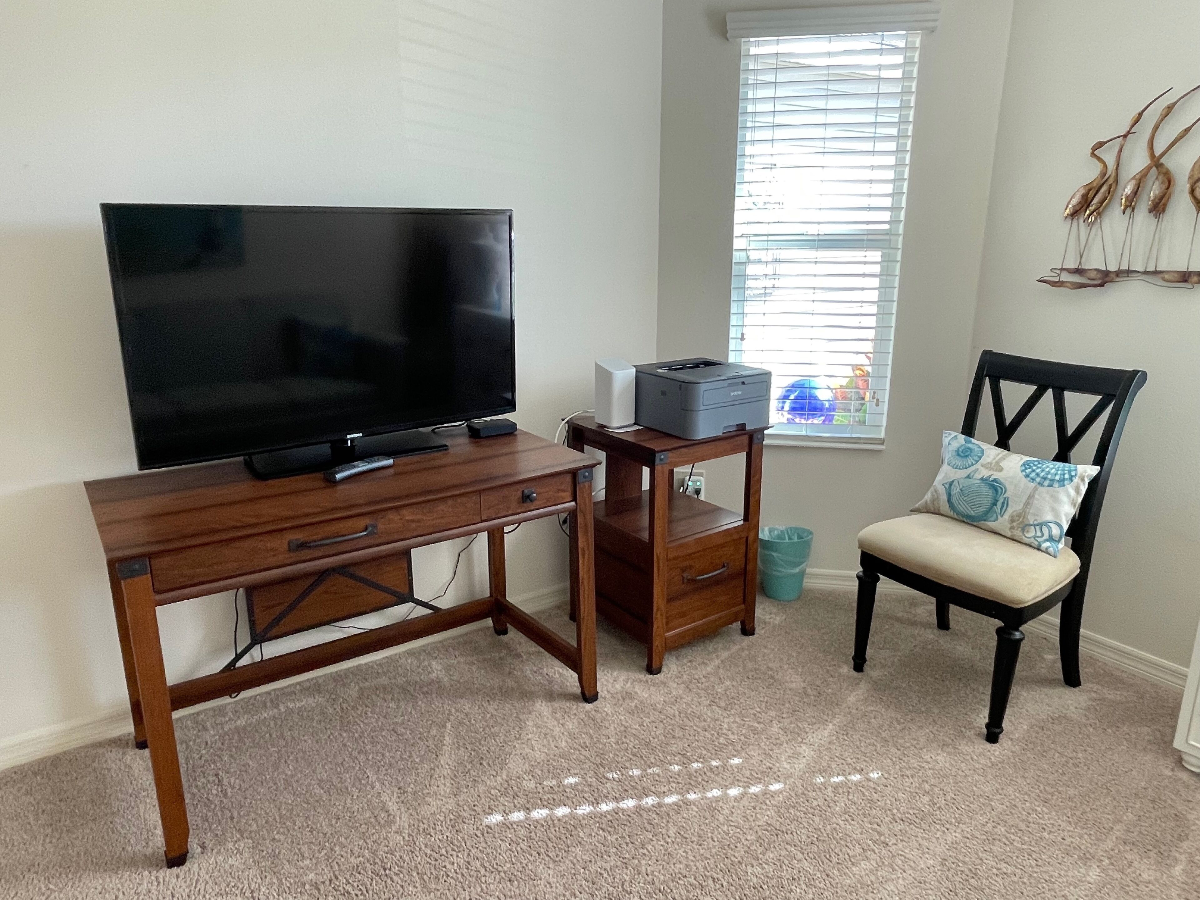 A room with a wooden desk, a television, a printer, a chair with a cushion, and a window with blinds.
