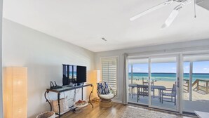 A room with a wooden floor, a TV on a stand, a chair, a table, and a view of the beach through a glass door.