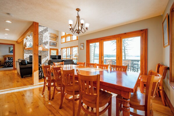A spacious dining area with a large wooden table and chairs, a stone fireplace, and large windows offering a view of the outdoors.