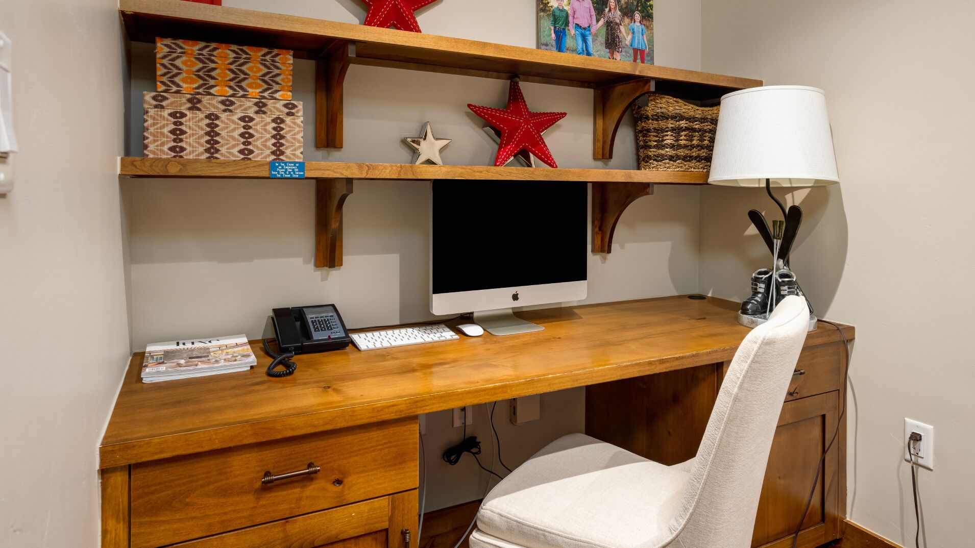 Un bureau en bois avec un ordinateur, un clavier et un téléphone. Des étagères se trouvent au-dessus, ornées d’objets décoratifs, et une lampe est placée à droite.