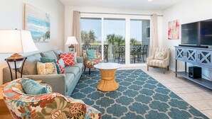 Ein Wohnzimmer mit einer Couch, Sesseln, einem Couchtisch, einem Fernseher und einem Balkon mit Gartenmöbeln.