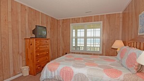 Quarto com painéis de madeira, cama com colcha florida, cômoda com televisão em cima, janela com persianas e mesa de cabeceira com abajur.