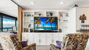 Sala de estar con un televisor de pantalla plana grande montado en un mueble blanco, rodeado de estanterías con objetos decorativos e imágenes enmarcadas. Hay dos sillones estampados y una mesita con una lámpara.
