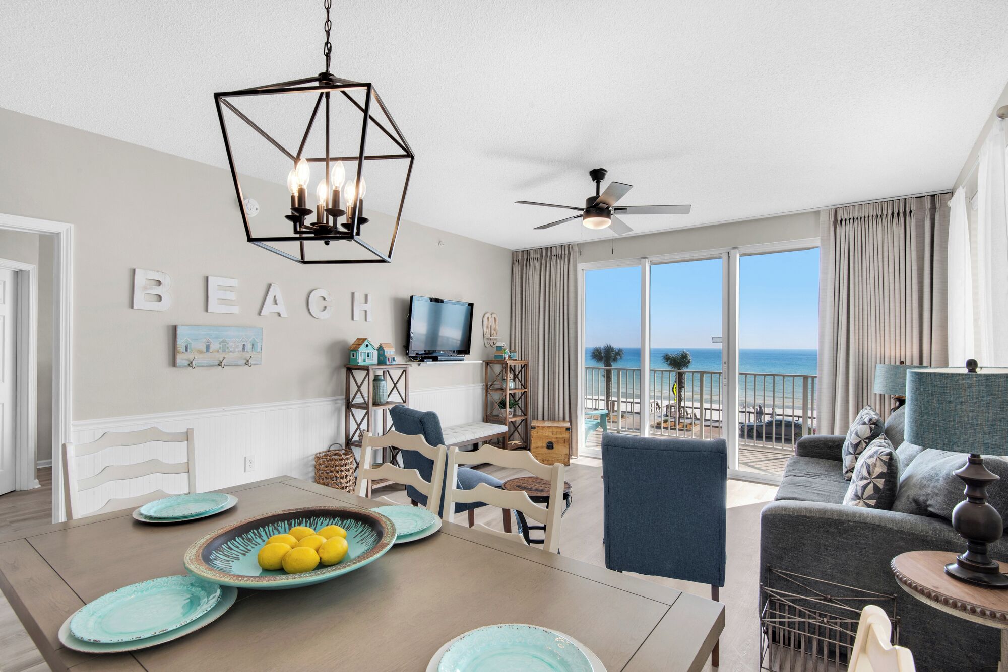 A modern living room with a dining area, a ceiling fan, a TV, and a view of the beach.