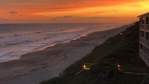 Vista costeira com praia, ondas do oceano e um edifício com varanda que dá para a paisagem.