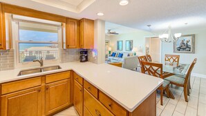 A modern kitchen with wooden cabinets, a large island, and a dining area with a glass table and chairs. - Beachside & Amazing Views! 3BR/3BA, Heated Pool/Spa  606 (Redington Shores)