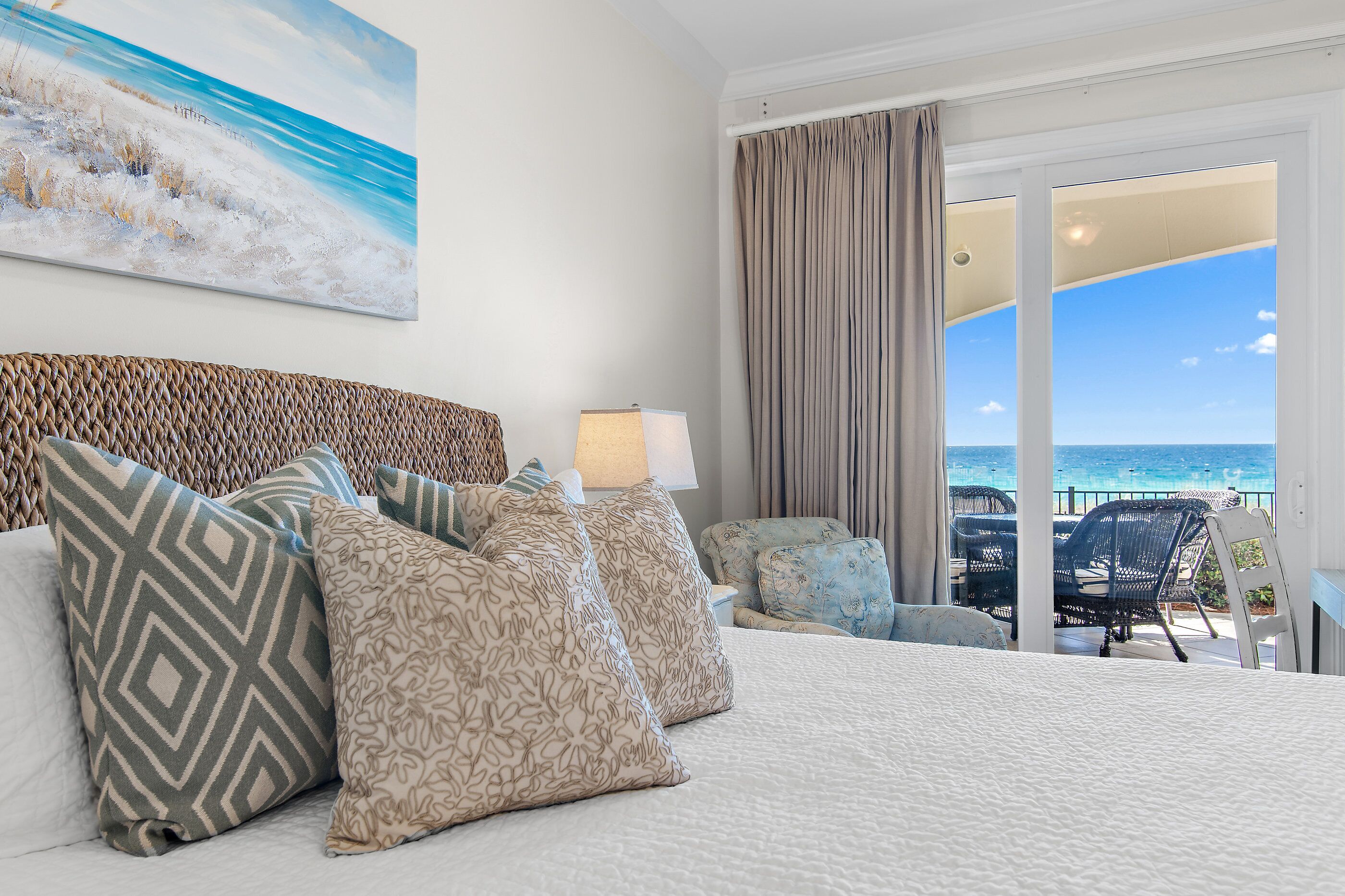 Ein Hotelzimmer mit Bett, Kissen, einer Lampe und Blick auf das Meer durch große Fenster.