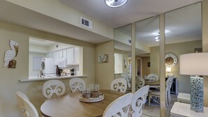 A dining area with a round table, white chairs, a kitchen in the background, and a large mirror reflecting the room. - 238 Shorewood- 2nd Floor Landscape View steps to the beach. (Hilton Head Island)