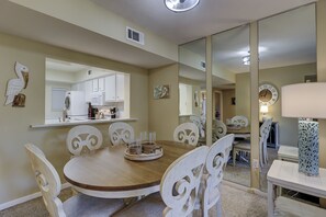 Uma sala de jantar com uma mesa redonda, cadeiras brancas, cozinha ao fundo e um espelho grande refletindo o ambiente.