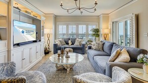 A spacious living room with a large flat-screen TV, a chandelier, and a comfortable seating area with sofas and a coffee table. - 3 bedroom/3 bathroom 2nd floor oceanfront condo with lots of amenities (Hilton Head Island)