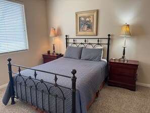 Une chambre à coucher avec un lit en métal, une table de chevet avec une lampe, une commode avec une lampe, un tableau encadré au mur et une fenêtre avec des stores.