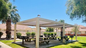 Un espace pique-nique abrité, doté de bancs et de tables à pique-nique, entouré de pelouse et de palmiers.