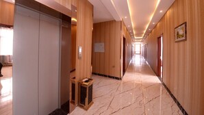 Corredor longo e bem iluminado, com painéis de madeira e piso de mármore, que leva a elevadores e portas.