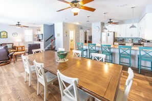 A spacious dining area with a wooden table and white chairs, a kitchen with white cabinets and a blue backsplash, and a ceiling fan.