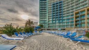 Ein Strand mit blauen Liegestühlen und Sonnenschirmen, ein hohes Hotelgebäude im Hintergrund und ein bewölkter Himmel.