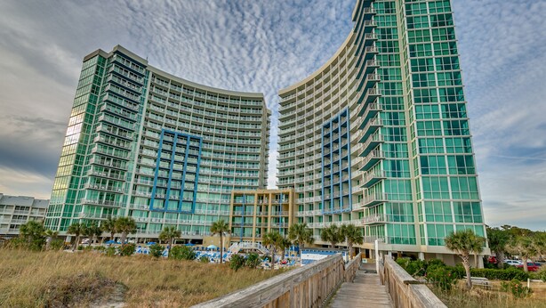 Ein Hochhotel mit gewölbter Fassade, mehreren Balkonen und einer Promenade, die zu einem Poolbereich führt.