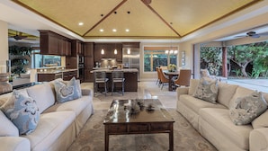 A spacious living area with a large sofa, coffee table, and decorative pillows. A kitchen with dark wooden cabinets and stainless steel appliances is visible in the background. - KaMilo 321 - Beautiful 3 bedroom with private pool! (Kamuela)