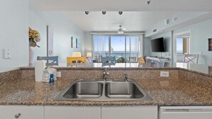 Una cocina con encimera de granito, pileta doble y vistas al área de estar, con ventilador de techo y grandes ventanales.