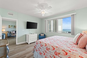 Quarto com cama, ventoinha no teto, televisão, janela com vista e porta para uma sala de estar.