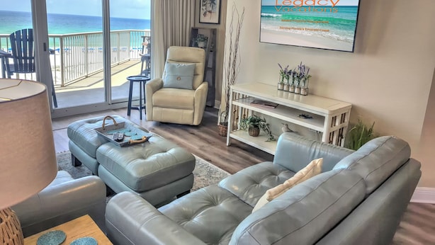 Tilava olohuone, jossa on suuri ikkuna, josta avautuu näkymä rannalle. Huoneessa on mukava sohva, pieni pöytä ja televisio, jolla näkyy ohjelma nimeltä ”Legacy Vacations”.