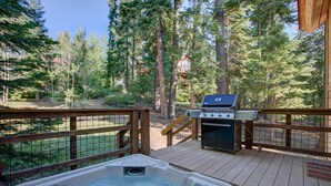 Una terraza de madera con jacuzzi, parrilla y vistas a un área boscosa.