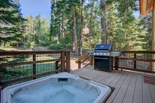 Eine Holzterrasse mit Whirlpool, Grill und Blick auf einen bewaldeten Bereich.