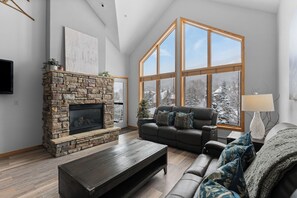 Un salon avec une cheminée en pierre, une grande fenêtre donnant sur des arbres couverts de neige, un canapé et une table basse.